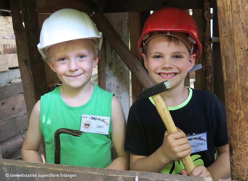 News-Bild Baustelle: Zwei Kinder in einer Holzhütte mit Bauhelmen auf dem Kopf und einem Hammer in der Hand